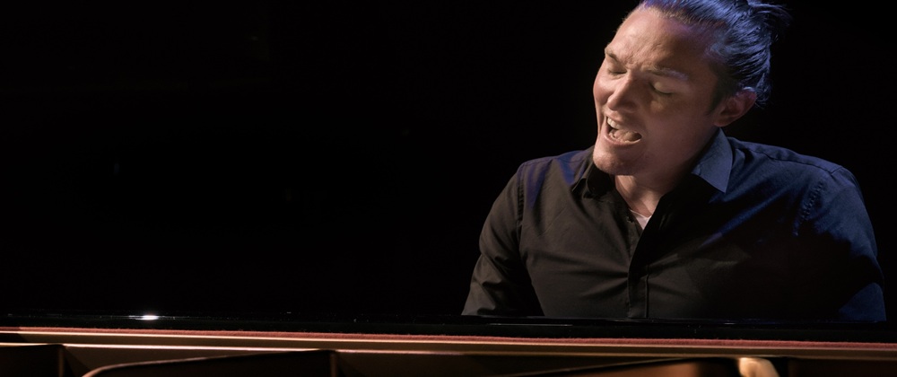 Andreas Langsch, Klavierkabarettist. Sitzt innig singend am Flügel vor schwarzem Hintergrund,